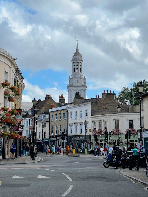 The image depicts a bustling town square with historic buildings and a prominent clock tower with a gold weather vane on top, set against a partly cloudy sky. The foreground shows pedestrians walking 