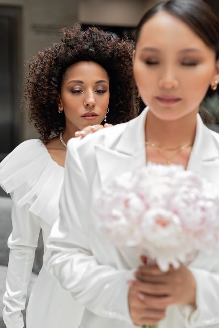 A close-up of a woman with straight dark hair and a white blazer holding a floral bouquet, with delicate pink and white roses, wrapped in white tissue paper. In the background, a woman with curly dark