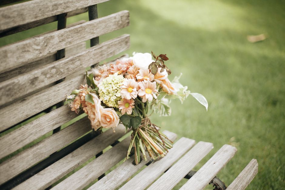 A bouquet of fresh flowers resting on a weathered wooden park bench outdoors. The arrangement includes peach and cream roses, light pink primroses with yellow centers, white hydrangeas, and greenery w