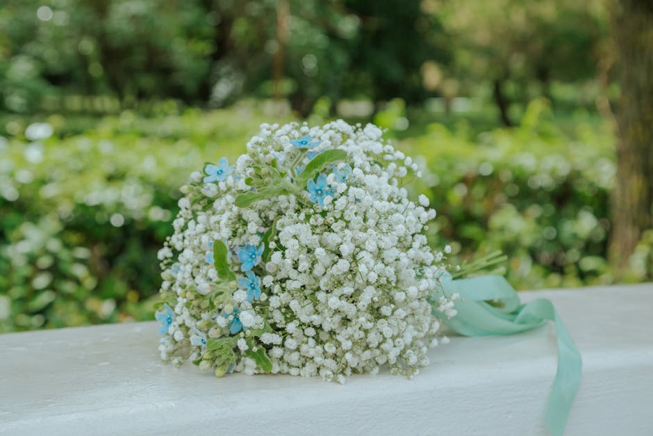 A delicate bouquet of fresh white baby's breath flowers, accented with small blue flowers and green foliage, arranged in a rounded, loose style. The bouquet is wrapped with a light blue ribbon tied in